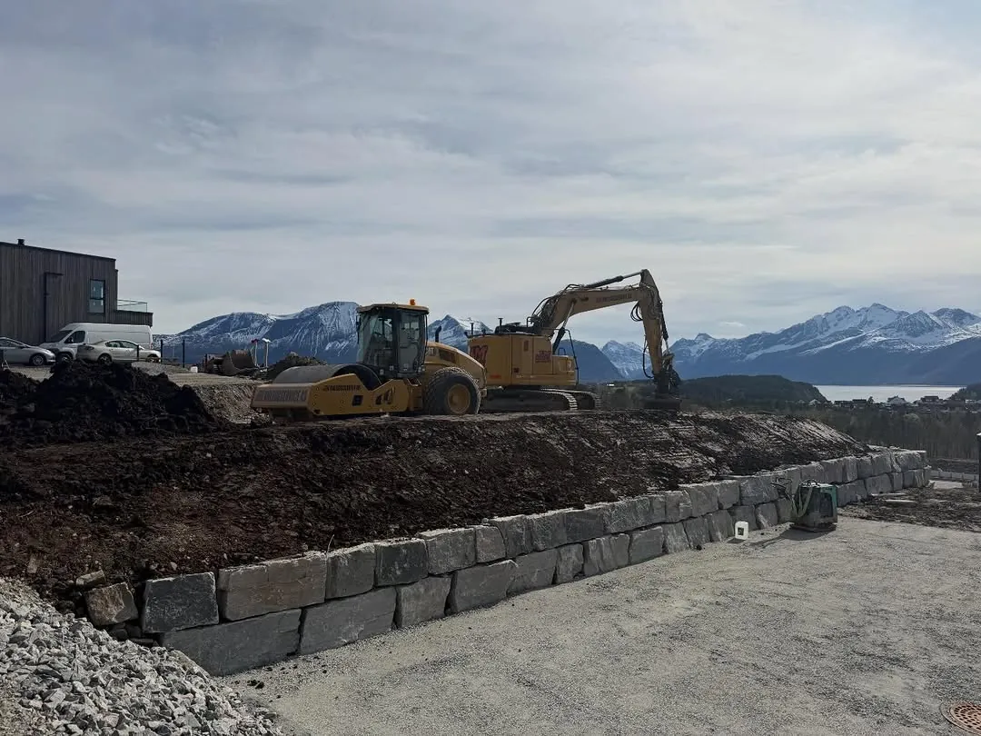 Gravemaskin ved en ny natursteinsmur, med snødekte fjell i bakgrunnen