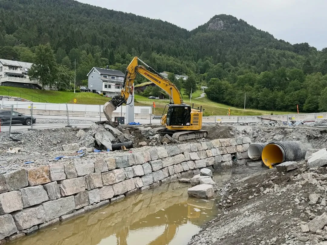 Natursteinsmur langs en bekk, med gravemaskin og stikkrenne